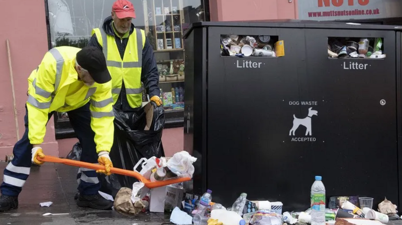 Two bin workers in yellow hi vis clothing use a shovel to collect rubbish from the street beside an overflowing black bin