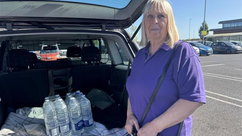 A woman with shoulder-length blonde hair and a purple polo shirt stands beside a car with an open boot, showing it contains half a dozen bottles of water.