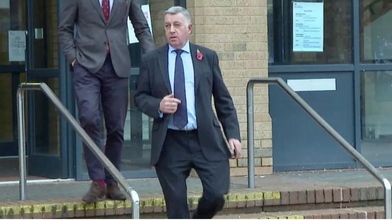 A man with short grey hair is leaving the magistrates' court. He is wearing a dark blue suit, with a light blue shirt and a dark blue tie. He has a poppy on his left lapel