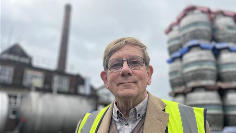 Miles Jenner who has glasses and a high vis jacket on. standing next to barrels of beer stacked up. And the harvey's building and large brick chimney behind him
