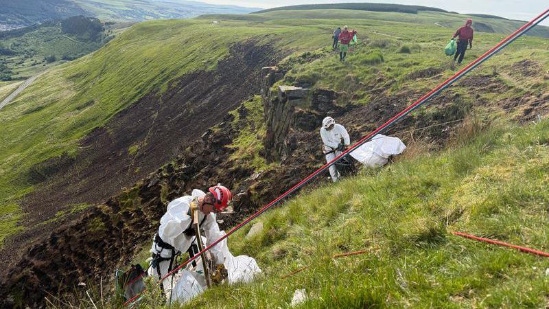 Dau wirfoddolwr mewn ofarôls gwyn a helmedau yn casglu sbwriel ar y mynydd