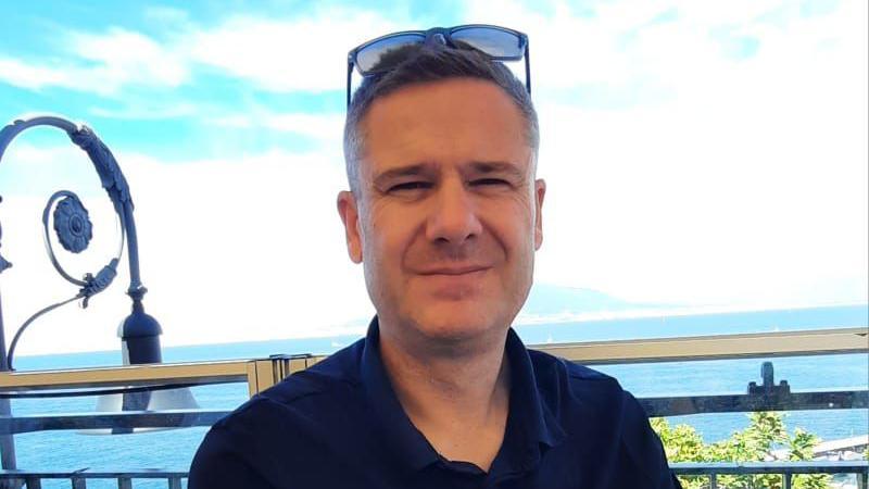 Andy Probert on holiday with the sea behind him on a hot, sunny day. He has short black hair with sunglasses perched on top of his head. He is wearing a black polo top with the top button undone and he is smiling while squinting.