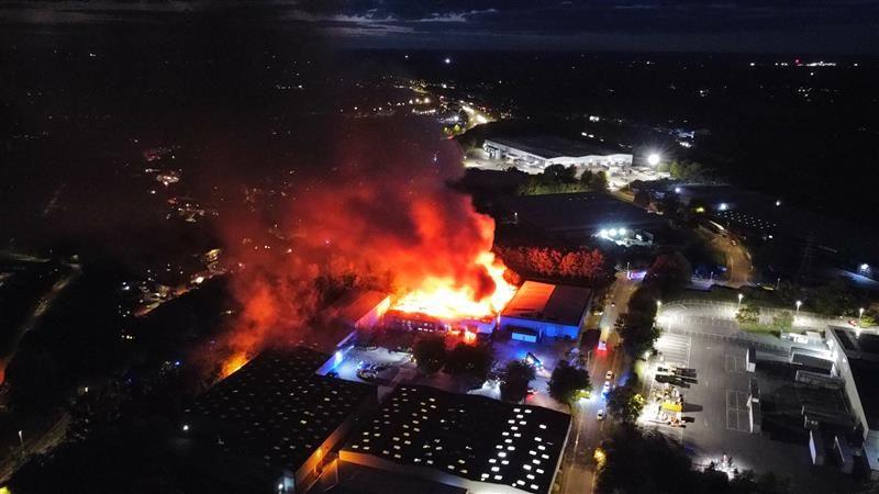 Drone footage shows a series of industrial units in the dark. In the middle is one unit on fire with red flames and smoke coming from the top
