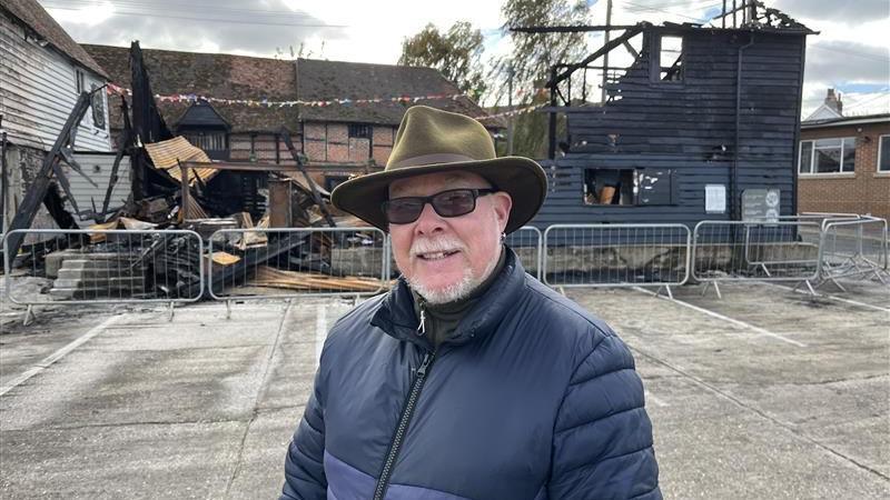 A man wearing sunglasses, an olive hat and a black puffer jacket. He is standing in front of a rubble of a fire-damaged building.