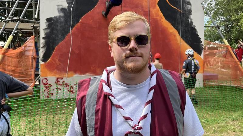 Ed Brackley wearing sunglasses, a white top and a red hi-viz jacket. Behind him is a wall with people climbing up it