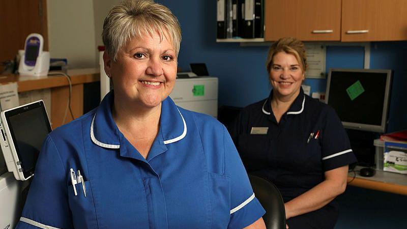 Two nurses smiling in a clinic - one in the foreground with a light blue uniform and one behind and too the right with a dark blue uniform