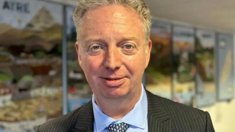 Alex Allinson, a man with light curly hair, looking directly at the camera. He is wearing a dark suit with a blue shirt and tie and there are blurred artworks featuring Manx landscapes on the wall behind him. 