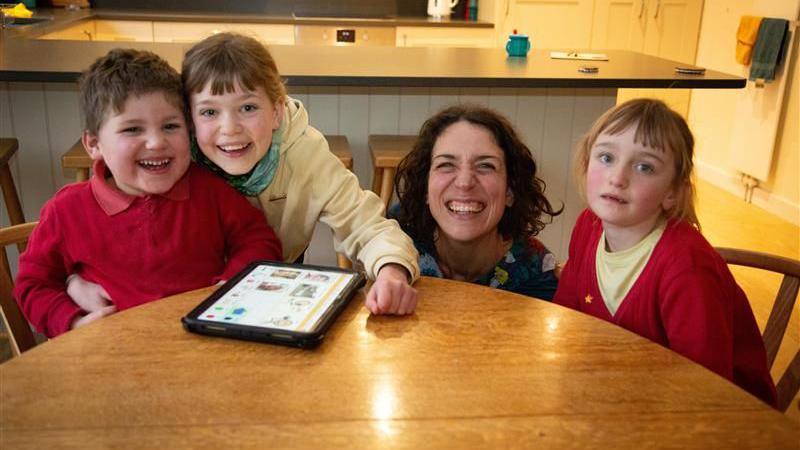 A five-year-old boy, two school-aged girls and their mum are sat around a dining table in their kitchen, smiling broadly at the camera. A tablet rests on the table. The boy has short brown hair and is wearing a red polo shirt and red sweatshirt. One of the girls, who has brown tied back hair and is wearing a yellow hoodie, has her arm around him. Next to her is their mum who is crouched down behind the table and has short, dark curly hair. On her other side is the other girl who is wearing a yellow t shirt and red cardigan and has brown tied-back hair.