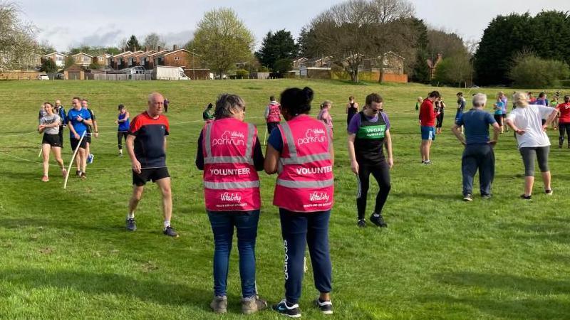 A group of runners on a grass park. One man in a red and blue top is approaching the finish, where there are two volunteers wearing parkrun vests