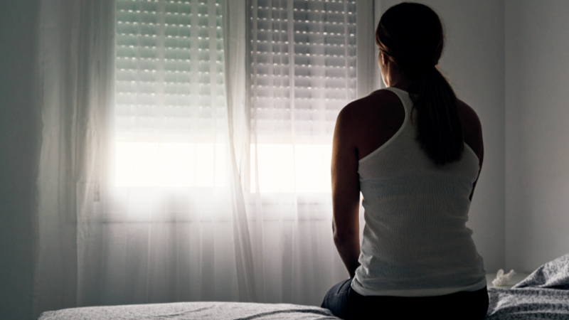 A woman sits on a bed with her back to the camera, looking towards a window