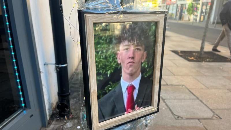 A framed photo of Thomas Taylor on a lamppost in Bedford near the bus station. He is wearing a black or navy blue suit, a red tie and has thick curly dark hair.