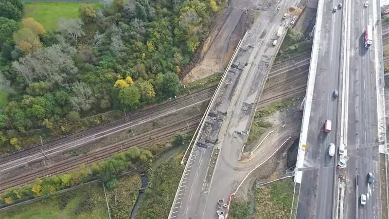 An aerial image of an old bridge, no longer in use with a new one next to it. Both cross a railway line.