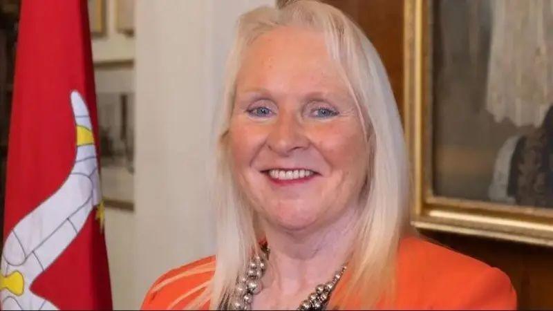 Julie Edge, a woman with blonde hair wearing an orange blazer smiling while standing next to a red Manx flag.