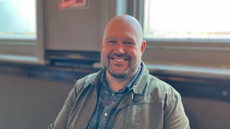 A bald man with a short beard wearing a khaki jacket