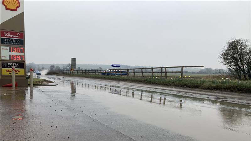 Looking down the road in the opposite direction. A petrol station is on the left. The road is streaked with ridges of water. In the distance a car hits a big puddle, splashing water in its wake.