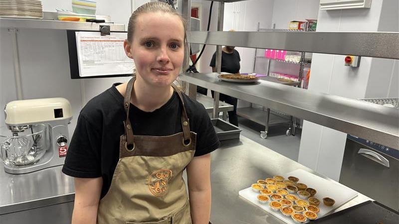 Picture of Georgia inside the café kitchen. She is wearing a beige apron and is stood at the side of a tray of canapés. 