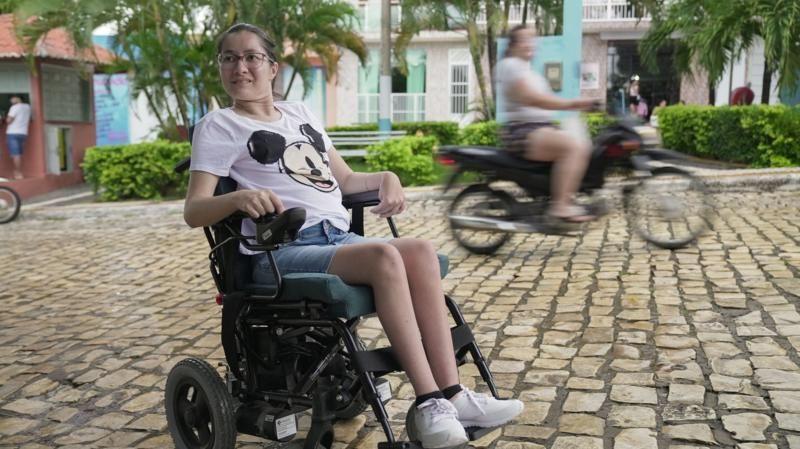 Paula, patient with Spoan, shown in a wheelchair on a cobbled street as a motorbike passes behind surrounded by bright green trees and bushes.