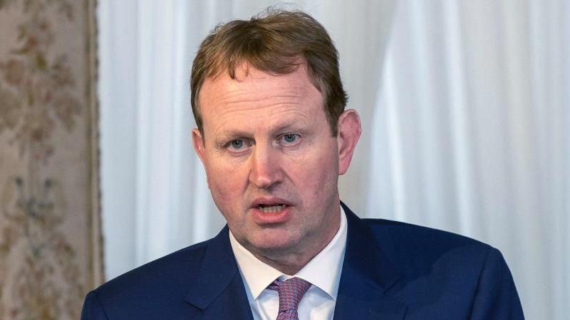 A man with short brown hair and blue eyes, wearing a blue navy suit jacket, white shirt and pink tie.