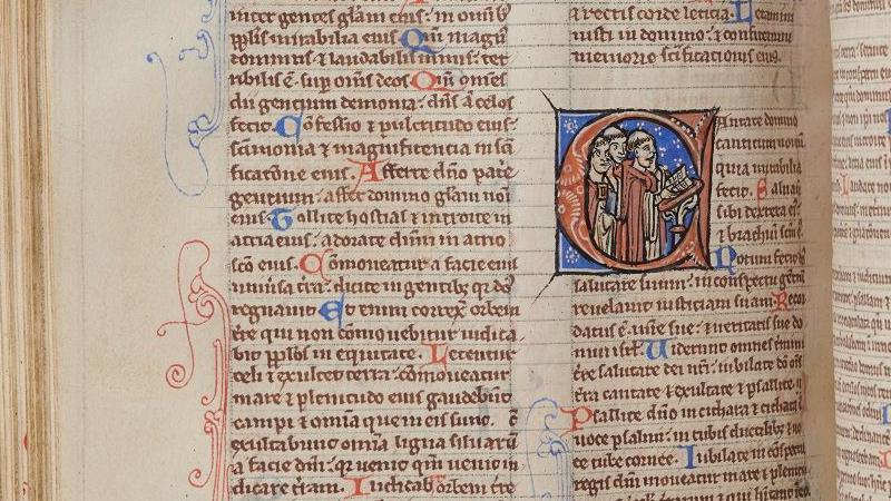 An old manuscript page, in tiny, perfectly written medieval Latin. One starting letter to a Bible book - a C - is lavishly decorated in blue and red and shows a group of men inside the letter reading from a lectern.