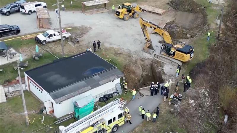 An aerial view shows emergency officials working around the sink hole, which is situated next to a one-storey building. A tractor, crane and fire engine are also nearby.