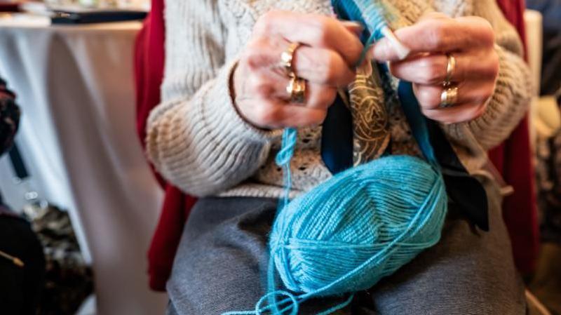 A woman's hands knitting with needles and a ball of light blue wool. She is wearing a cream jumper and a grey skirt.