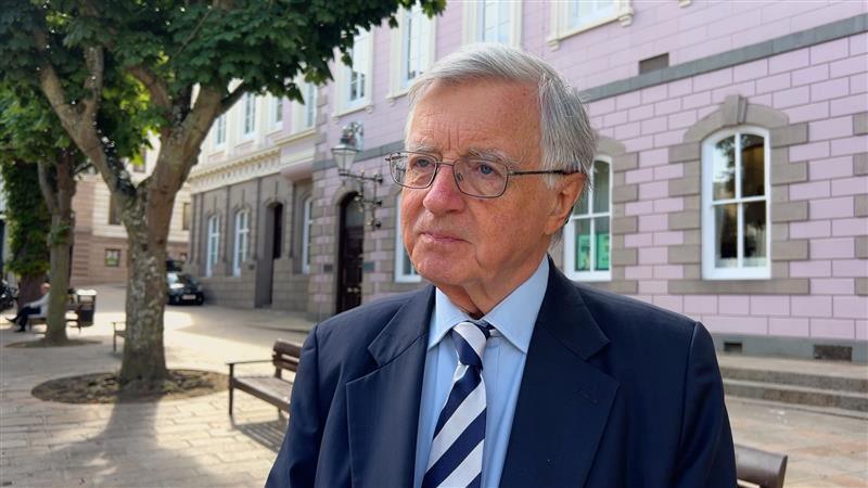 Deputy Sir Philip Bailhache wearing a blue suit and striped tie looks off to the left of the camera.