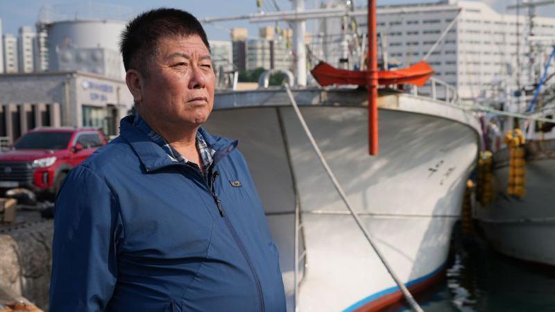 An older man in a blue jacket stands in front of a white fishing boat.