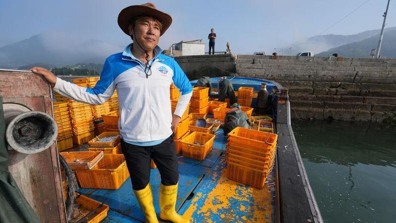 A man wearing a hat, a light jacket and bright yellow boots stands on the deck of a moored boat, alongside lots of yellow crates.