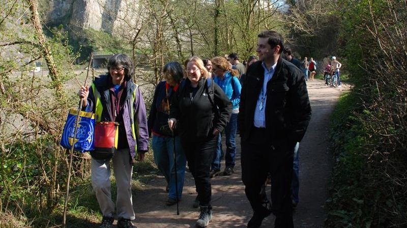 A group of people walking along a wooded path on a sunny day. One lady on the left is carrying a blue bag for life and a wooden stick.