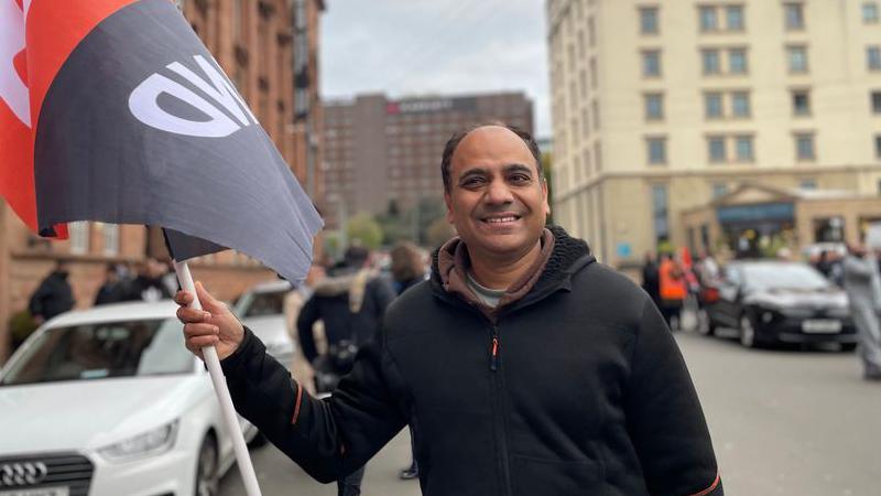 Vini Sharma, a balding man in a black top, smiles at the camera while holding a GMB flag