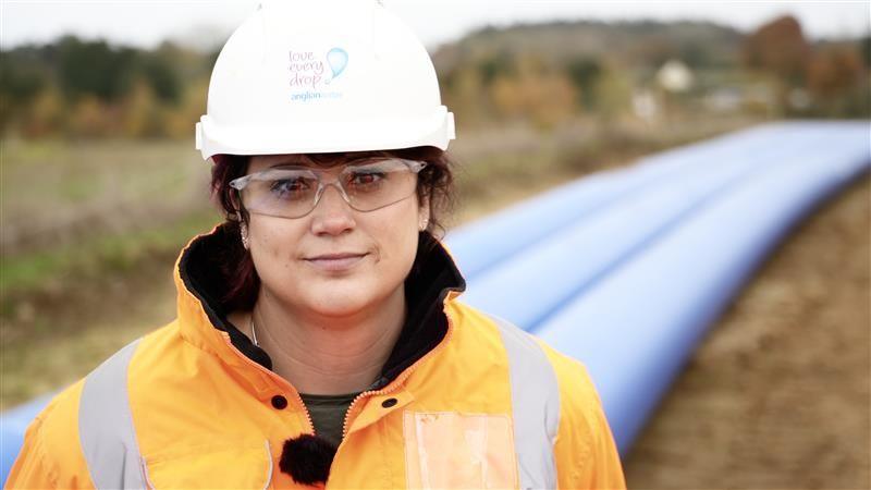 Regan Harris smiles at the camera in front of three blue water pipes. She is wearing a white coloured hard hat and an orange hi-vis coat. She is also wearing clear safety glasses. Her dark hair has been tucked into the hard hat.