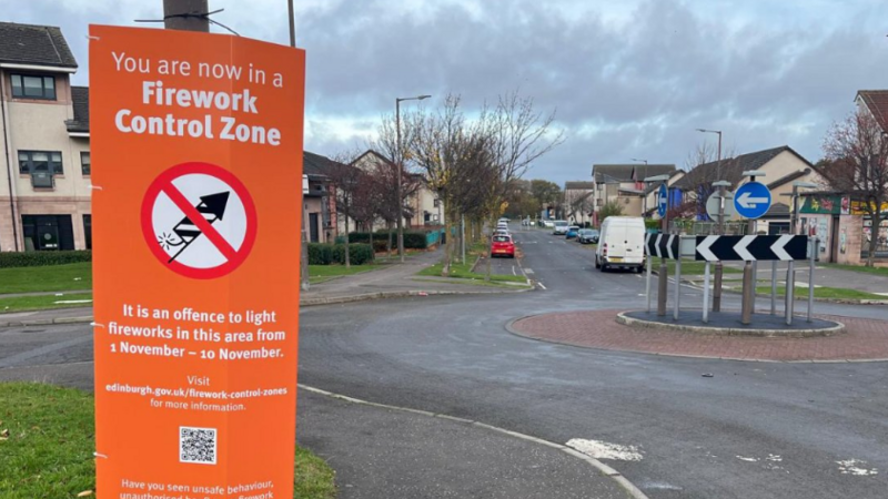 A large orange sign saying You are now in a firework control zone, with an illustration of a firework with a banned sign over it. The sign is in front of a roundabout and several houses.