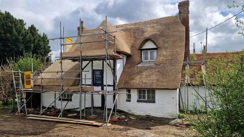 A house with a thatched roof. There are metal poles around it as it is being worked on. It is a cloudy day.