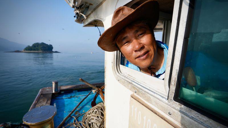 A man wearing a hat sticks his head out of the window of a boat. In the background is sea and an island.