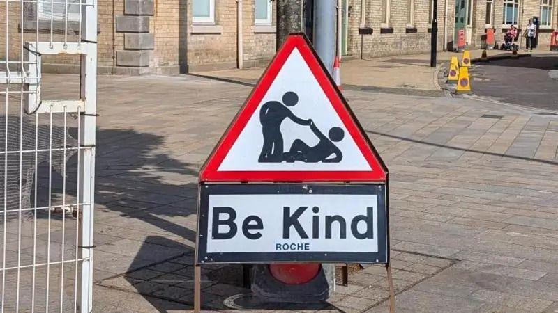 A triangular roadworks sign which normally features a man digging has been changed to show a man helping up another from the ground, with the words "Be Kind" replacing the word "Roadworks". 