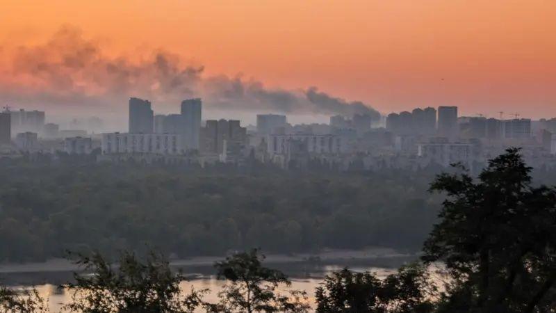 The skyline of Kyiv is stark and grey against an orange-hued sunrise, with smoke billowing into the sky after a recent strike.