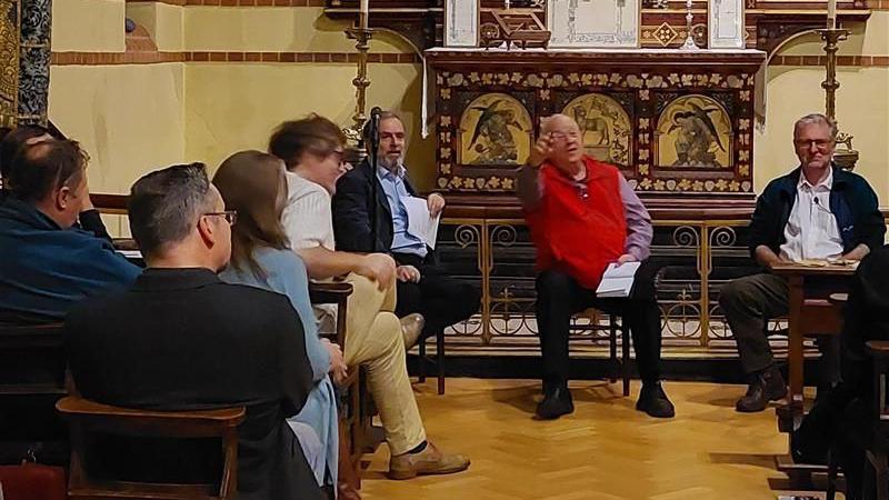 Audience members ask questions to Peter Hitchens and Richard Scarse. People are sitting at pews in front of the panel. Moderator John Mair has his right arm raised, pointing at a resident who has a question.