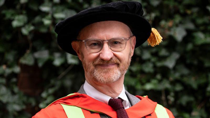 Mark Gatiss in a red and green graduation gown and a black velvet graduation tam hat.