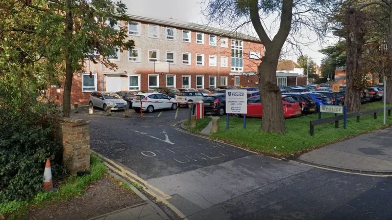 A general view of Slough police station, a three-storey building with 10 windows across. There is a car park outside it, with about 30 cars parked outside it, with an access road onto a larger road 