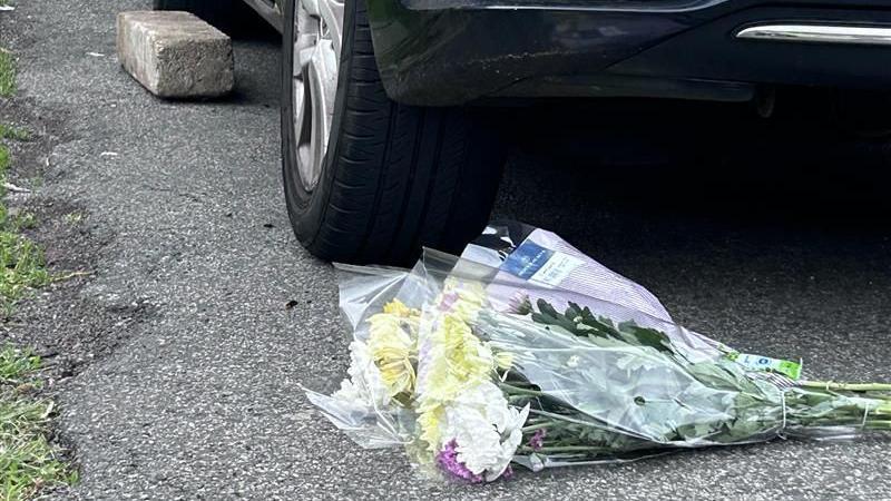 Two flower bouquets in plastic wrapping have been laid on a driveway in front of a car. The car's left back wheel can be seen in the picture.