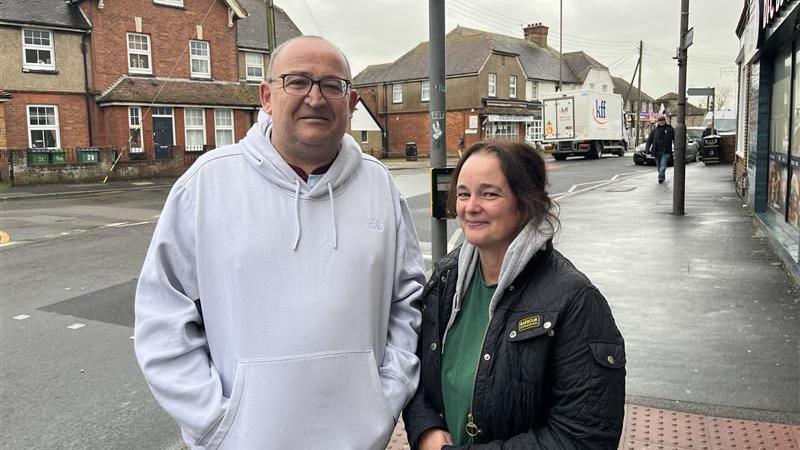 A man in a grey hoodie and a woman in a green top and black jacket, stood next to each other on a pavement. The road and some houses can be seen behind them.