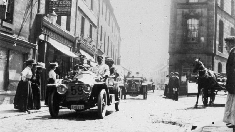 A black and white image of lots of very old racing cars going down a cobbled street with a group of women watching from the pavement 