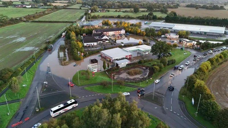 A drone view of buildings and a petrol station surrounded by floodwater. The buildings are adjacent to a roundabout. There is one car submerged in flood water, and a queue of traffic building to the right hand side of the site