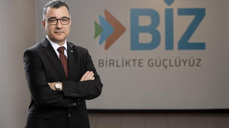 A man is wearing a suit and is standing with his arms crossed in front of a business logo. The logo says BIZ and then has some writing printed in Turkish. The man is smiling, has dark hair and is wearing glasses. 