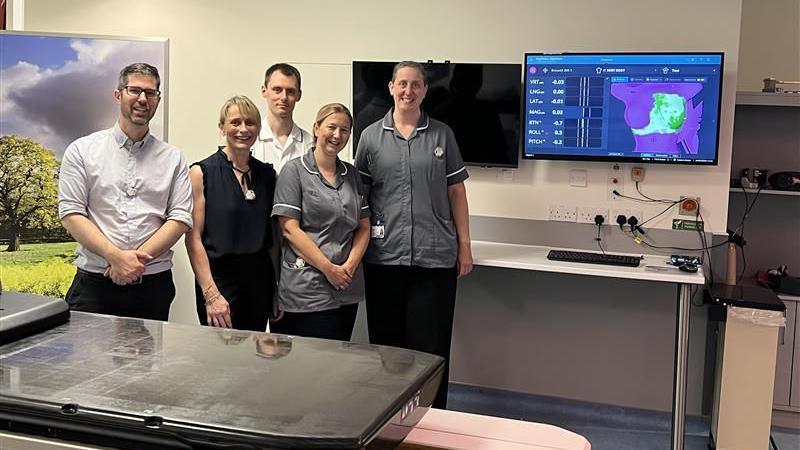 Five members of the hospital's radiotherapy team smliing at the camera standing beside a medical table and a computer screen