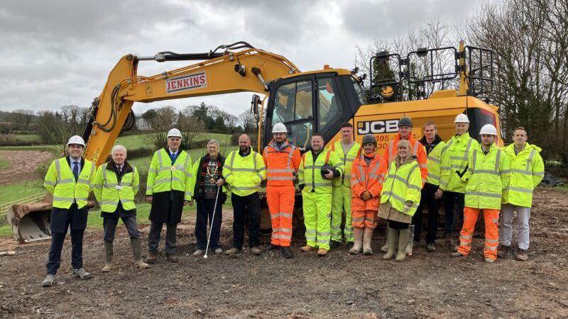 A group of 15 people pictured in front of a yellow JCB digger on a plot of land. The people are wearing high viz jackets and some are wearing hart hats.