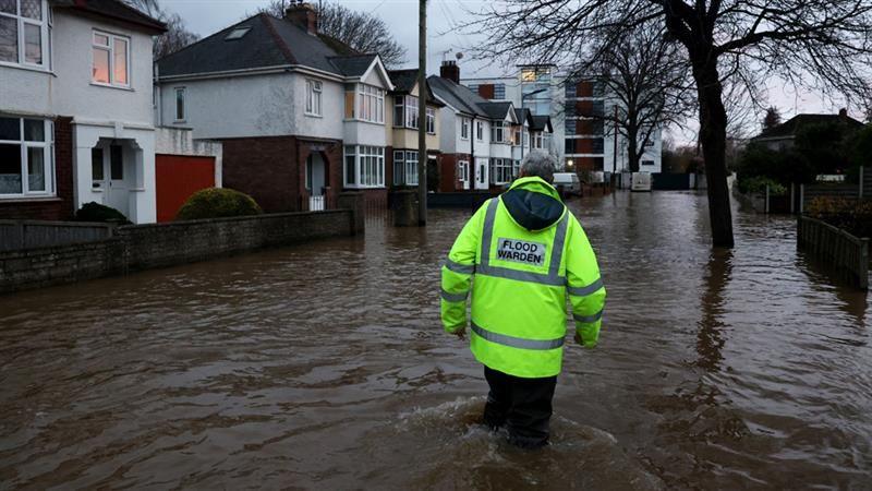 A man wearing a neon jacket with the words "Flood warden" on the back walks in knee-deep water beside homes