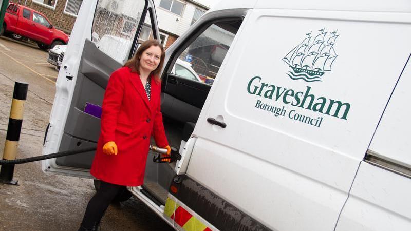 Councillor Emma Morley standing next to a white van. She is filling the car with a hose, and wearing a red coat and orange gloves