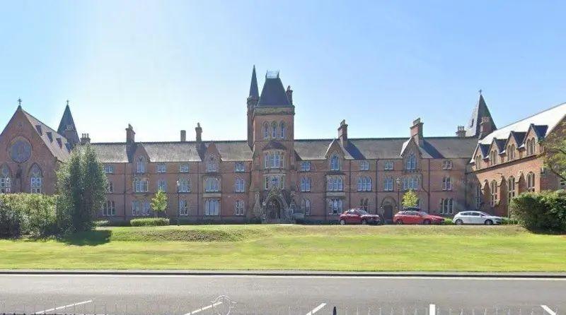 A large brick building sits on top of a lawn of green grass. The sky above is clear and blue. At the bottom of the frame is small black iron fencing. In front of the building there are two red cars and a white car.