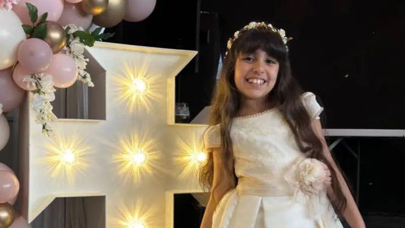 Alice da Silva Aguiar has long dark hair and is wearing a cream communion dress and floral headband. She is stood next to a white cross which is lit up and surrounded by pink, white and gold balloons